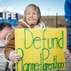 An older woman holds a yellow sign that says "Defund Planned Parenthood" in green letters, with a mountain in the background and people standing beside her