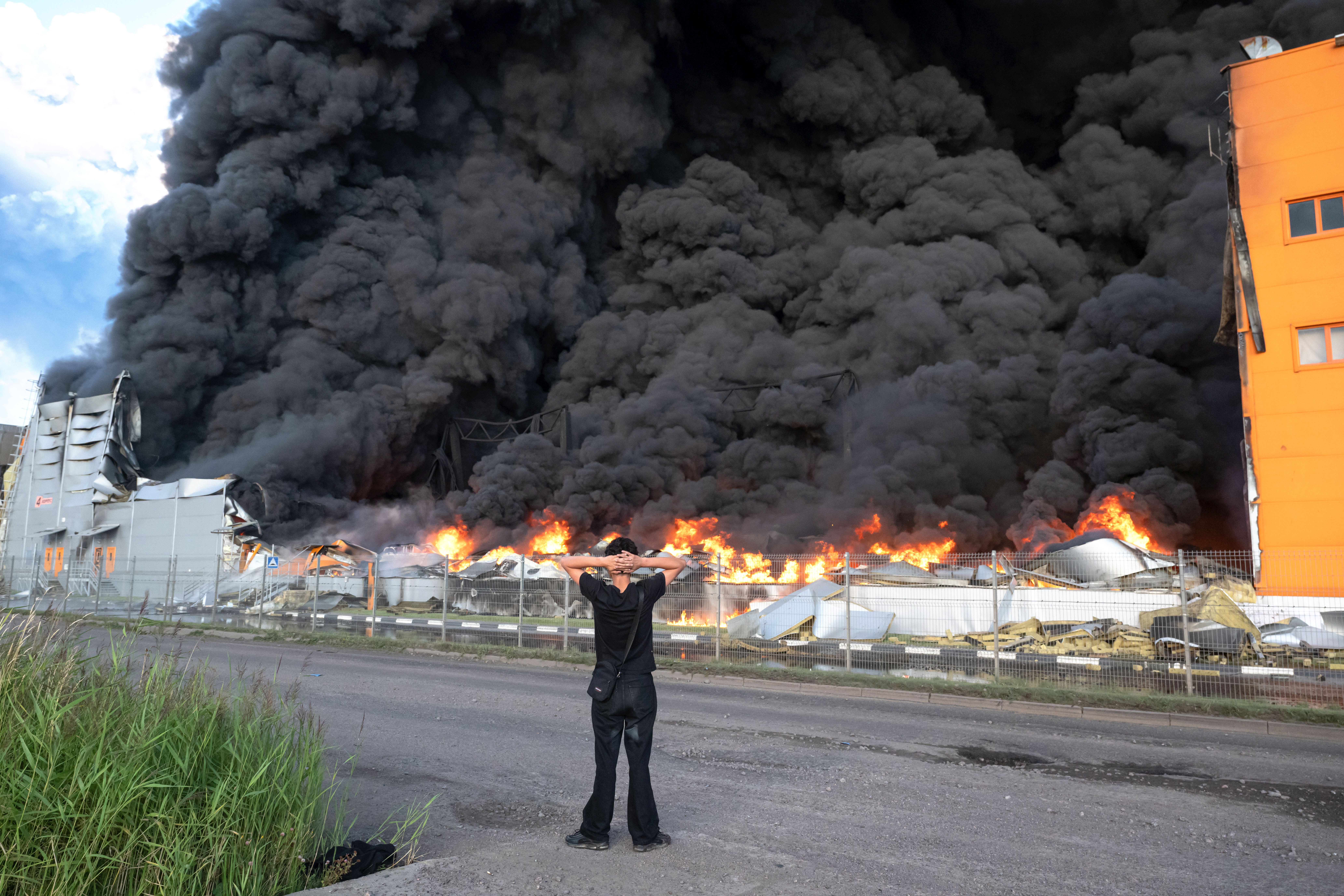 A man stands on a roadside, looking at at thick black smoke rising from a burning warehouse.