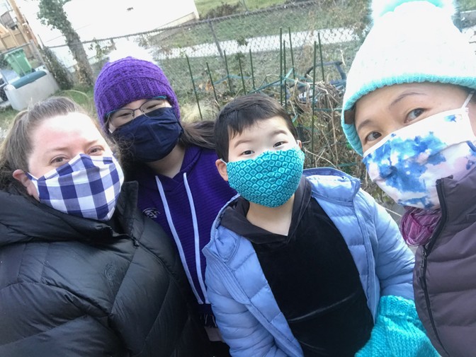 Two women and two young girls, wearing face masks and winter coats, gather outside in a yard