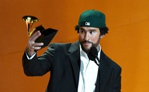Bad Bunny, wearing a white button-up shirt, a dark suit jacket, and a green baseball cap turned backwards, stands in front of a microphone holding a Grammy award.