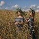 Two women wearing large wreaths of flowers stand in a wheat field.
