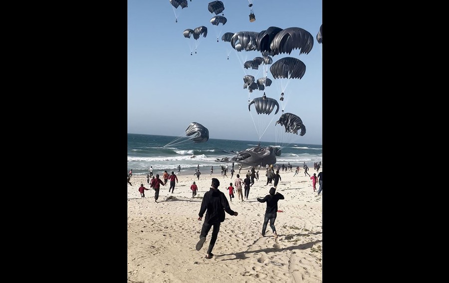 People run on a beach toward parachutes carrying aid packages falling into the surf.