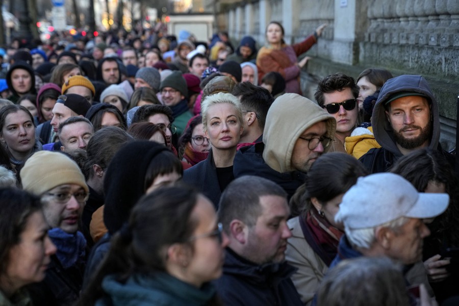 A woman stands in a crowd waiting outside a building, looking toward the camera.