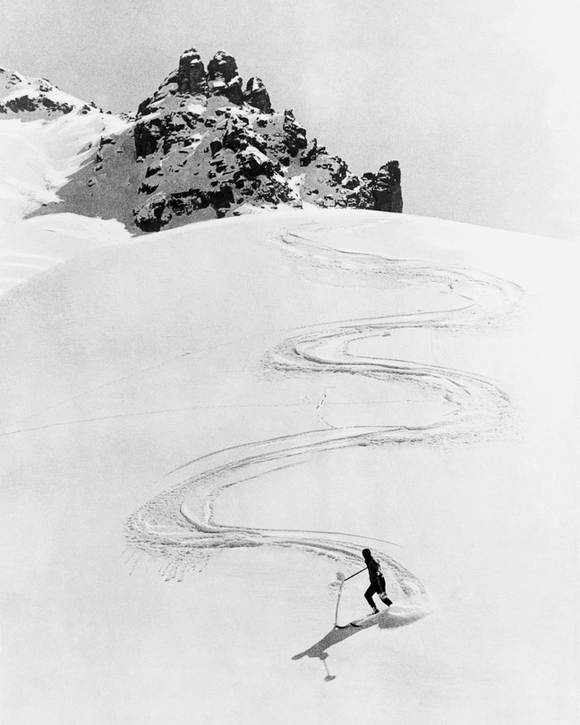 A black and white photograph of a skier coming down a steep alpine with a zig zag-pattern in the snow behind them