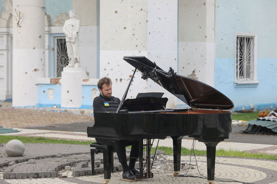 A person wearing a bulletproof vest plays a piano outside a pockmarked building.