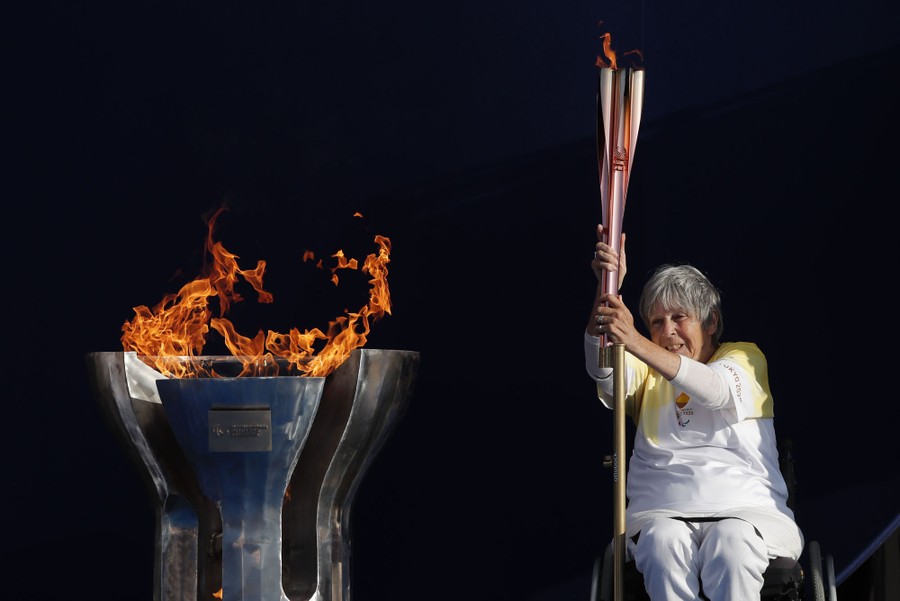 A woman in a wheelchair holds a lit torch beside a metal cauldron with flames rising above it.
