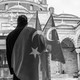 A black-and-white photo taken from behind a man looking at a mosque while holding two Turkish flags in his right hand.