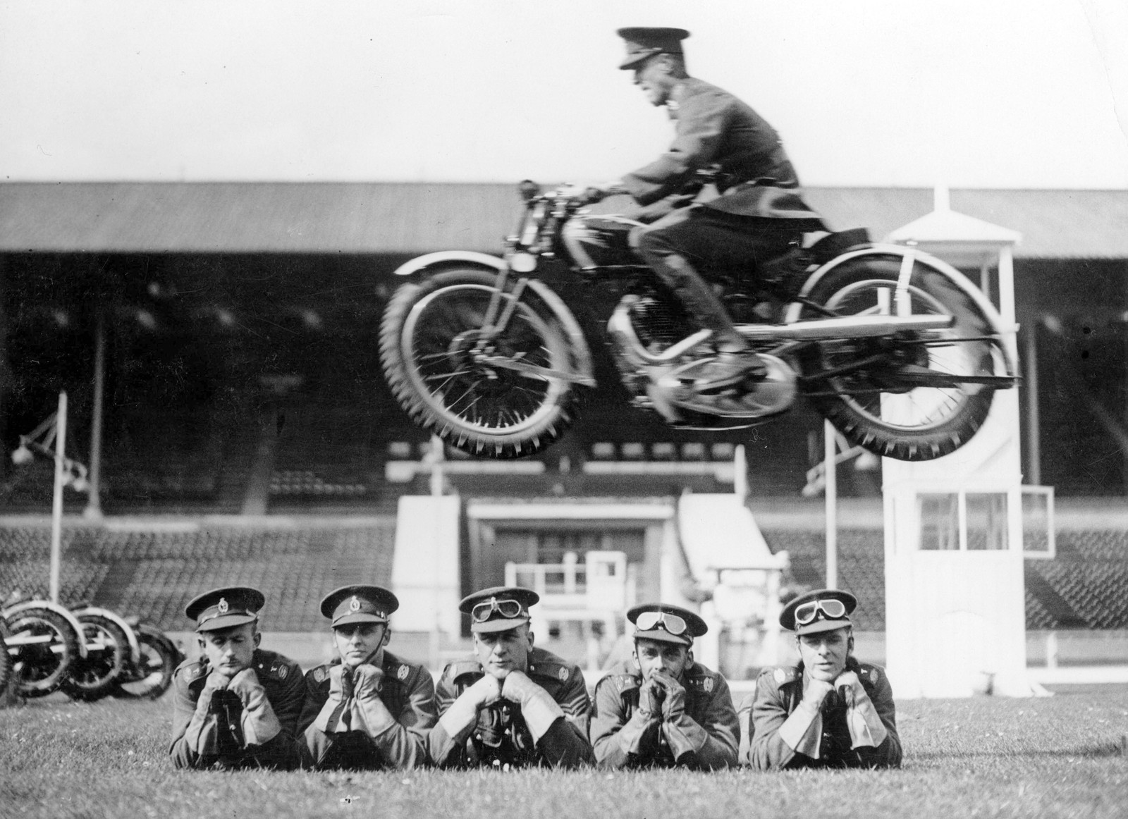 A soldier rides on a motorcycle, jumping over several other soldiers who are lying on the ground, facing the camera.