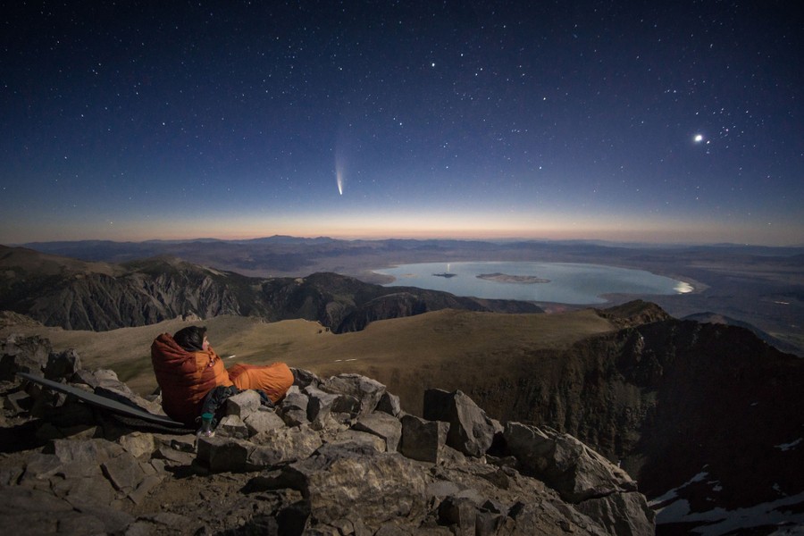 A person in a sleeping bag sits on rocks on a mountaintop, looking toward the horizon and the view of a comet in the night sky.