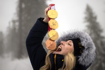 An athlete in a warm coat stands outside for a photo shoot, holding four Paralympic medals above their upturned face, pretending as if they might swallow them.
