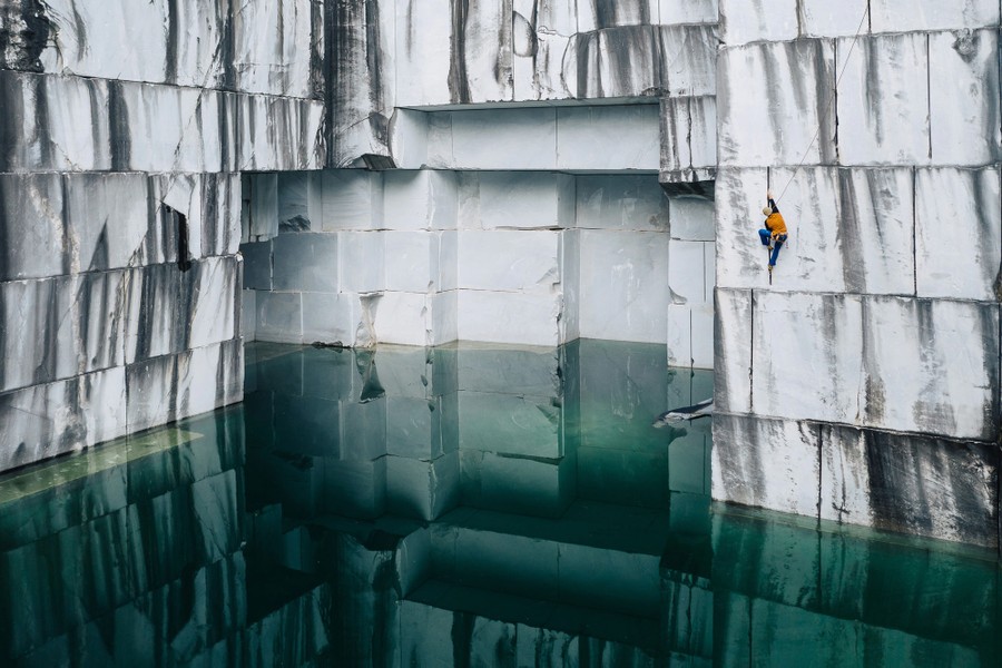 A climber hangs from cracks in the white walls of a marble quarry, above blue-green water.