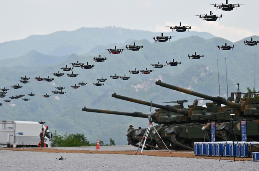 About 50 drones hover in formation in front of several tracked military vehicles.
