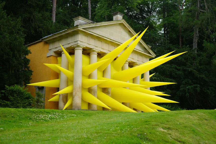 Giant yellow spikes, part of an inflatable art installation, poke out of columns of a building in a park.