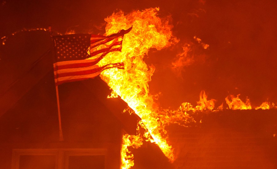 An American flag can be seen on a house that is burning in a forest fire.