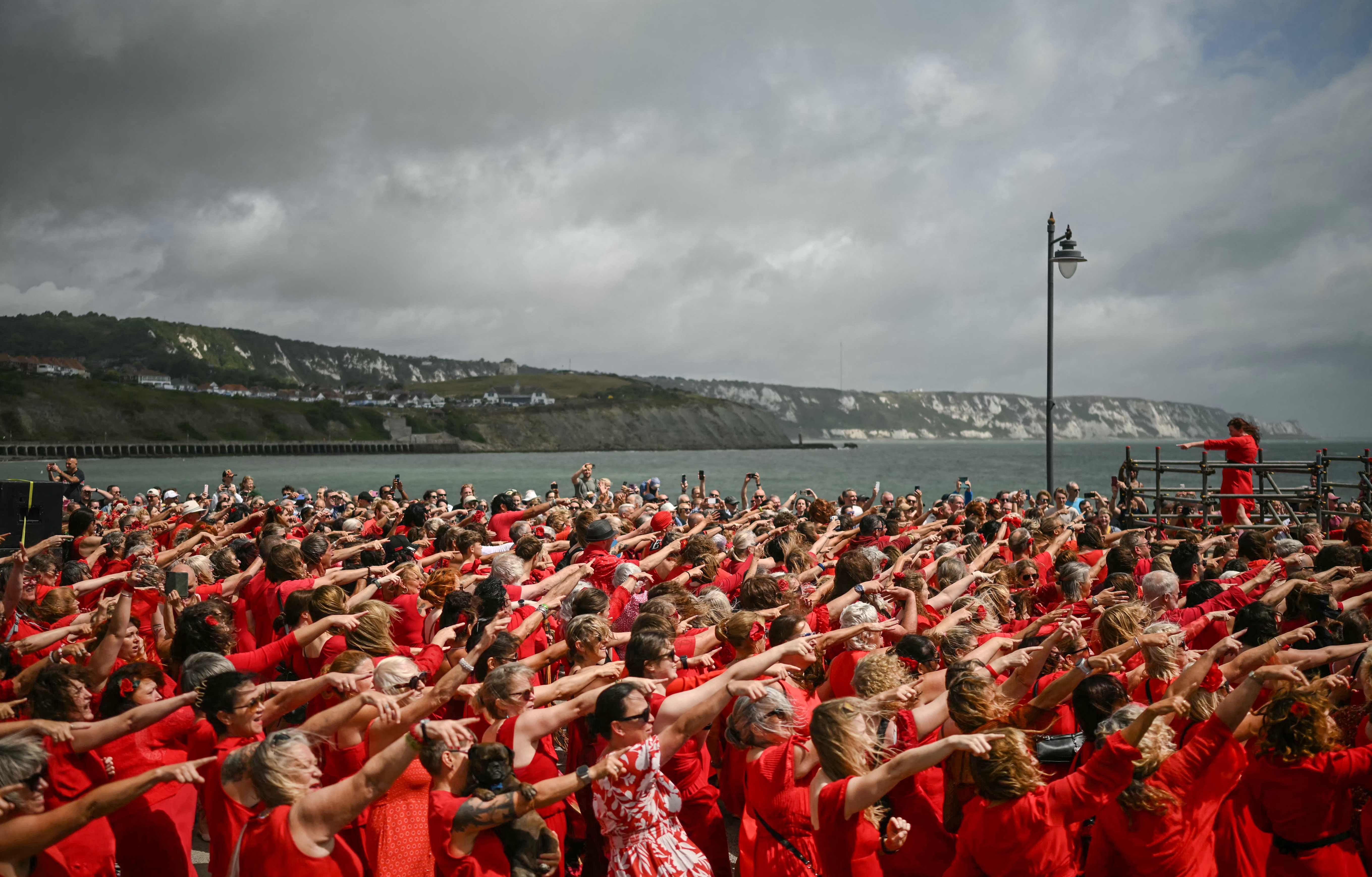 Hundreds of people dressed in red take part in a dance event along a coastline.