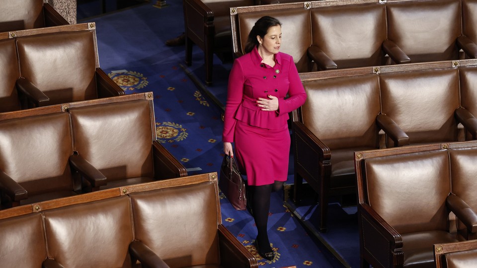 Elise Stefanik wears a pink suit and walks in front of empty seats in the House Chamber