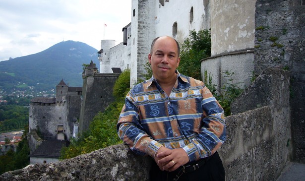 Michael Jones leans against a stone wall, smiling slightly, with a mountain behind him