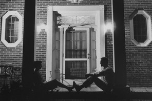 Black-and-white photo of a man and a woman sitting in front of a building at night. Each is leaning against opposite pillars, facing each other, with their feet touching.