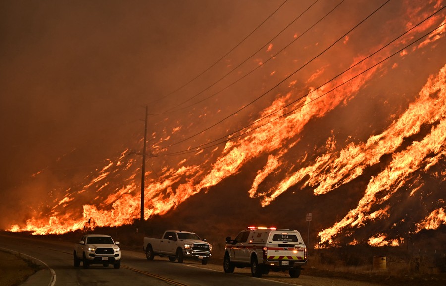 Three emergency vehicles sit on the side of the road as flames from a wildfire race up a hill.