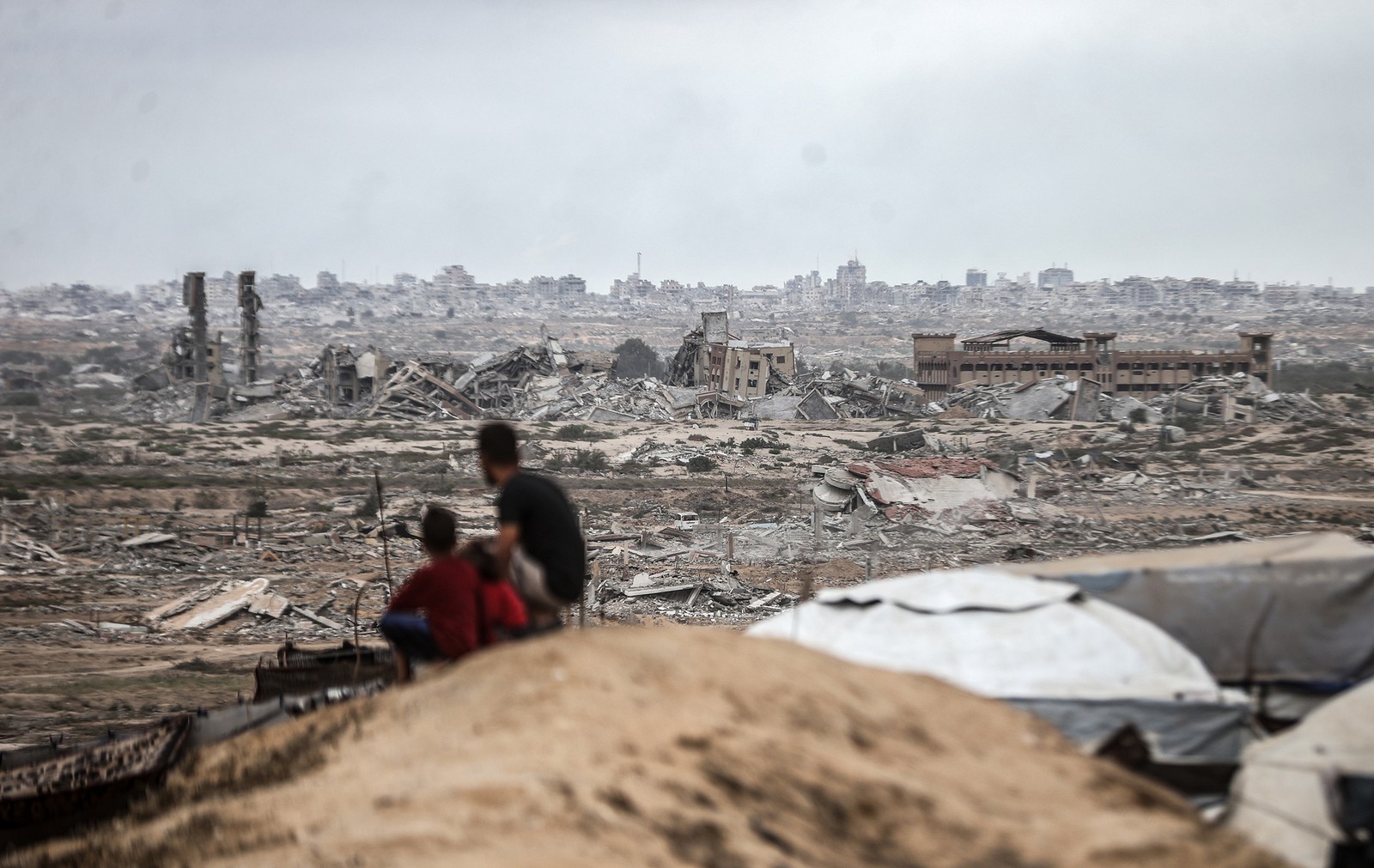 Several people sit on a barren hillside, looking out over a landscape of rubble and destroyed houses and buildings in Gaza.