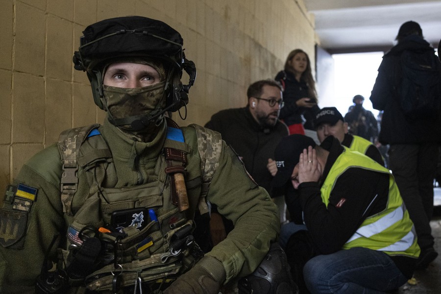 A soldier, several rescue workers, and civilians crouch together in a shelter.