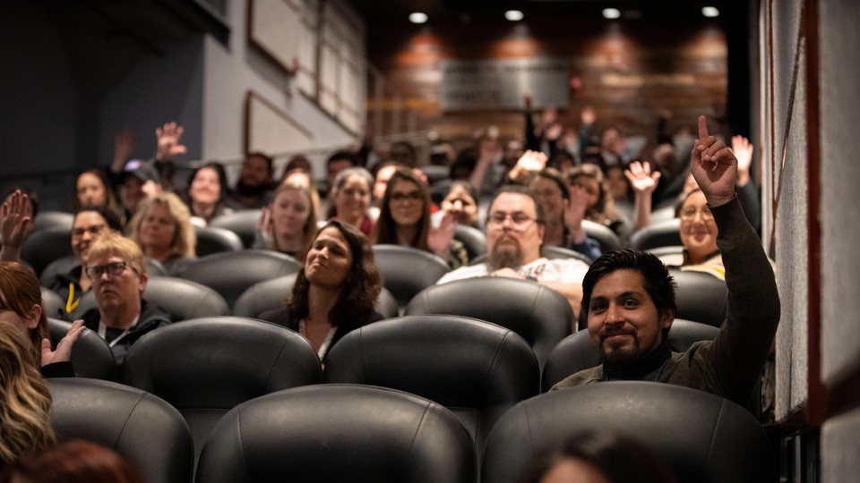 People are seated in an auditorium with rows of black leather seats. A few people raise their hands.