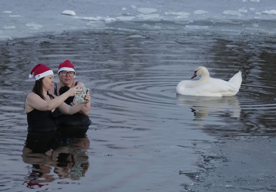 Two women in Santa hats and swimsuits take a selfie while standing in waist-deep lake water, near ice and a swan.