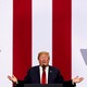 A photograph of Donald Trump standing behind a microphone and a pair of Teleprompters, in front of red-and-white-striped background