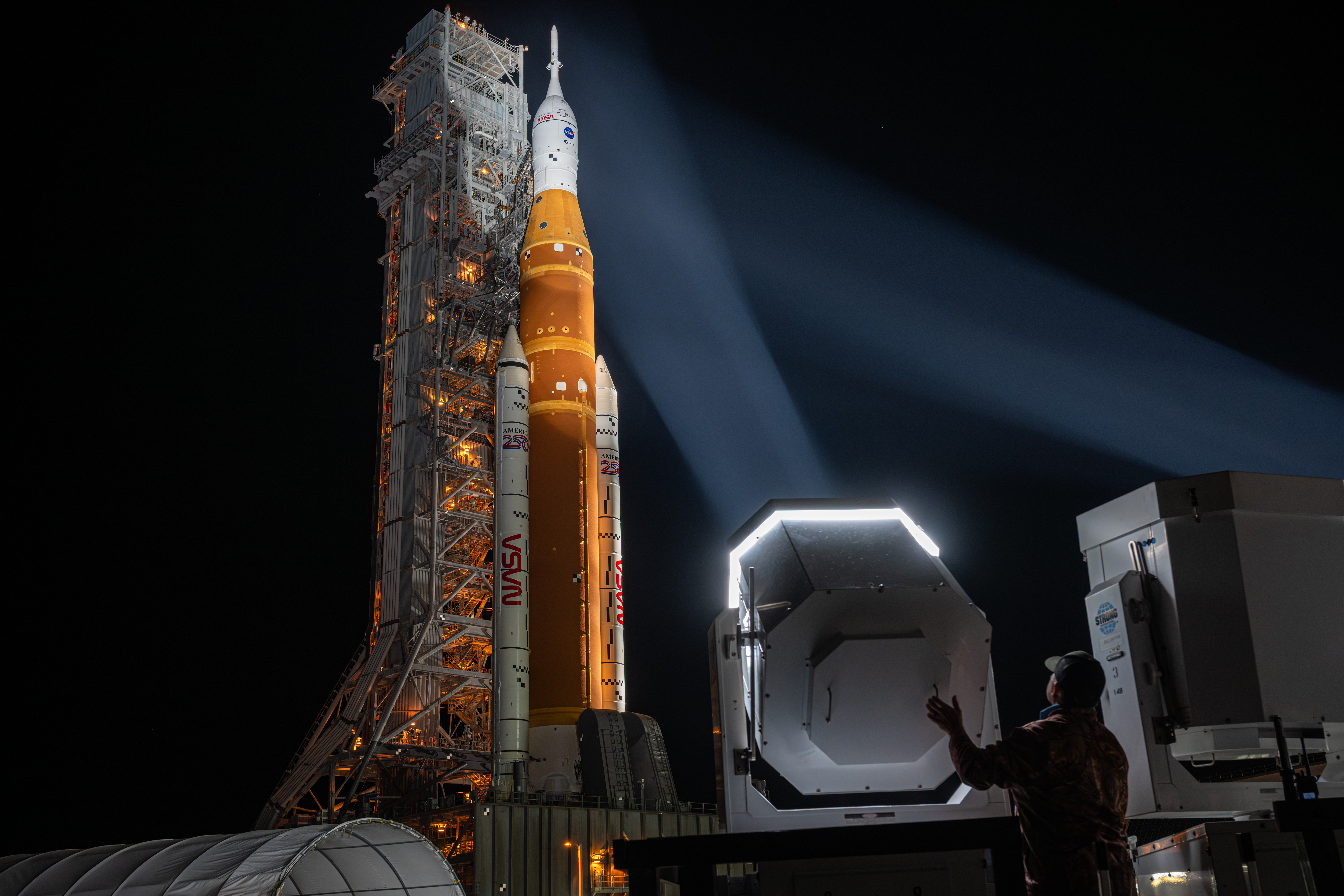 A person stands behind a spotlight that is aimed up at a tall rocket that is being rolled out to a launch pad.