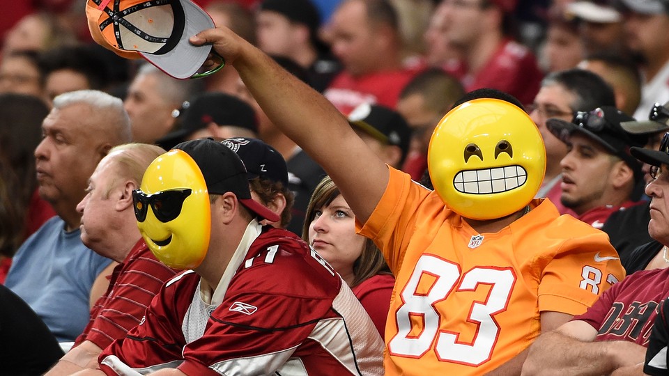 Fans wearing emoji masks wave from the stands at an NFL game.