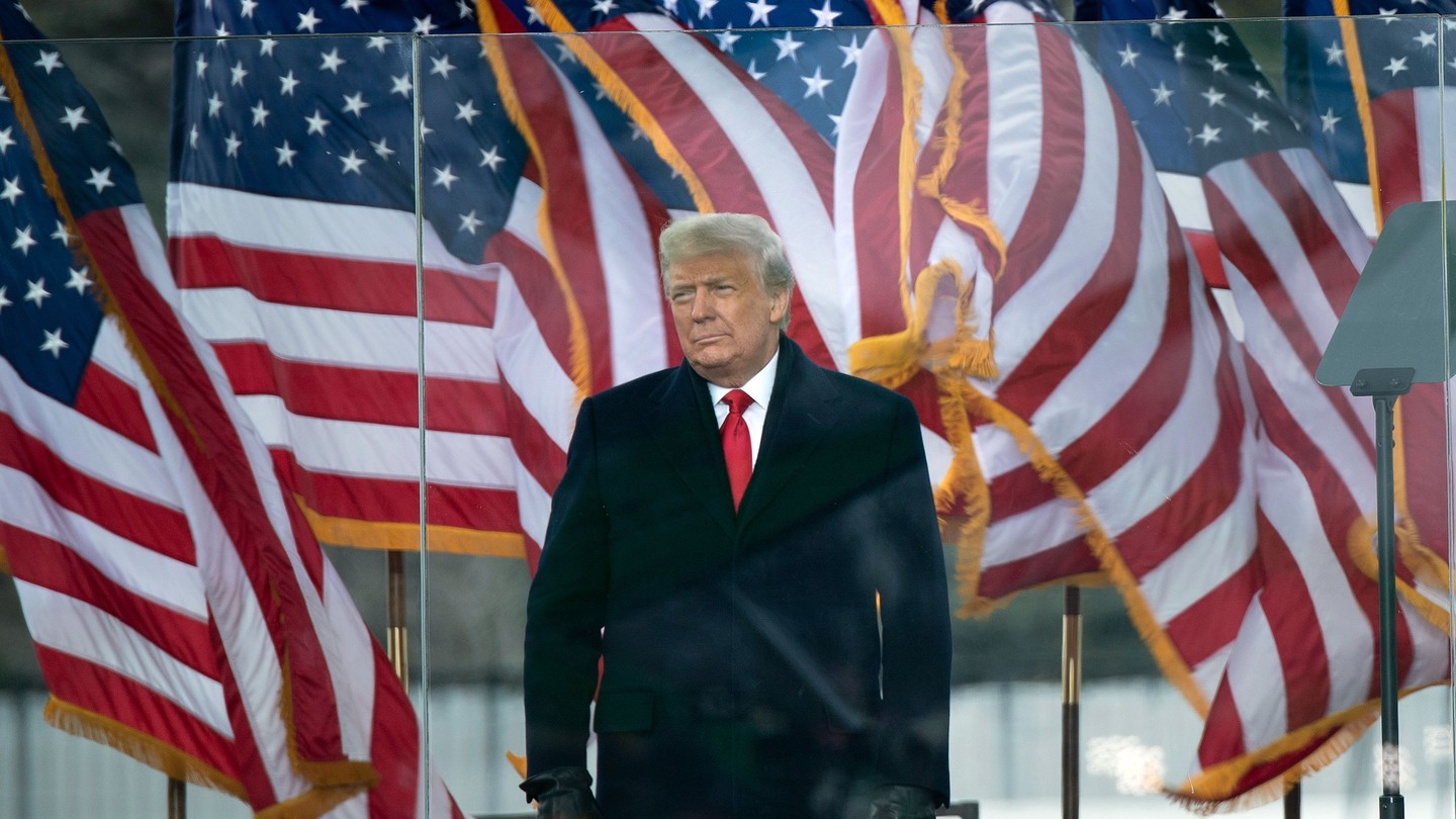 Donald Trump standing outside, behind clear barriers and in front of a row of billowing American flags