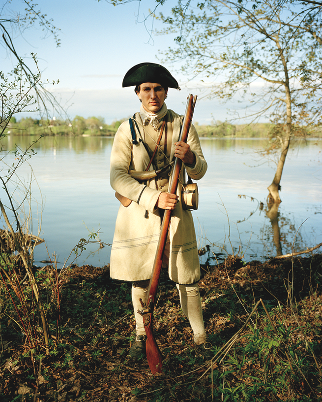 photo of reenactor as American soldier standing in black hat and tan coat with musket on the shore of Lake Champlain