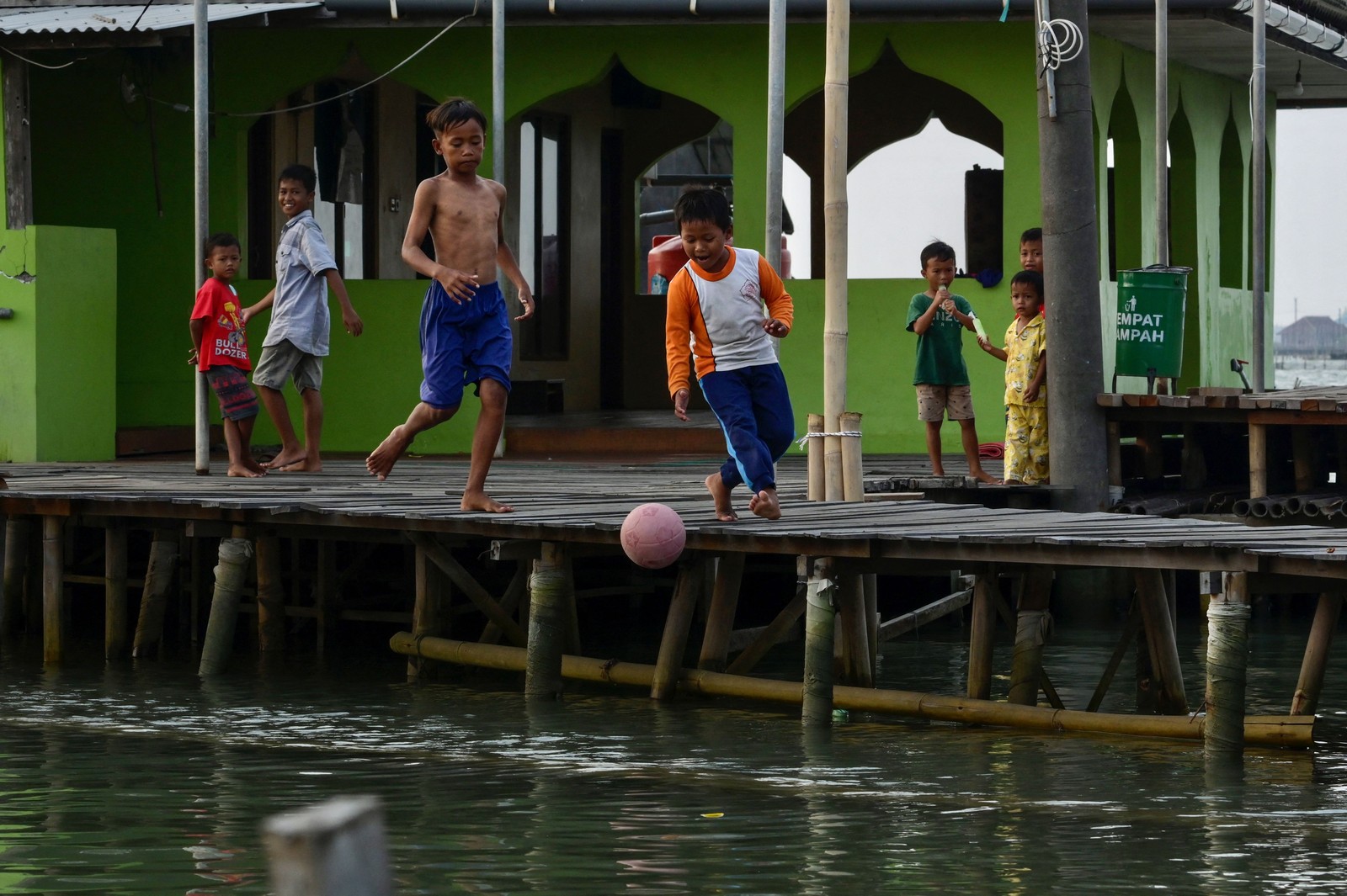 Several children play football on a raised boardwalk above seawater. The ball appears to have gotten away from them and is falling into the water.