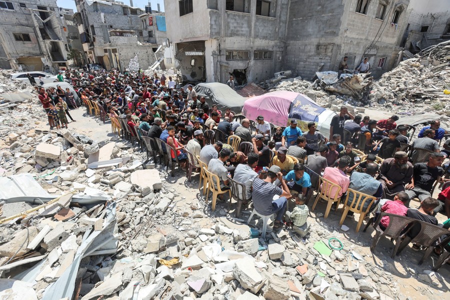 Dozens of people sit side by side in chairs set up on a road through the rubble of destroyed buildings.