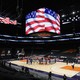 A basketball arena  with the players standing in a circle around center court and video screens showing the American flag above empty stands