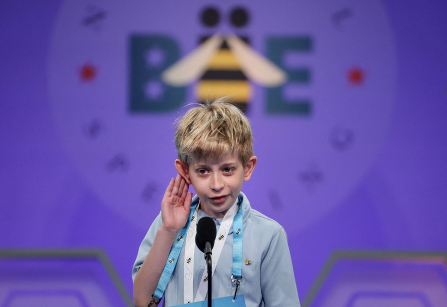 A young boy holds one hand to his ear while standing before a microphone during a spelling bee.