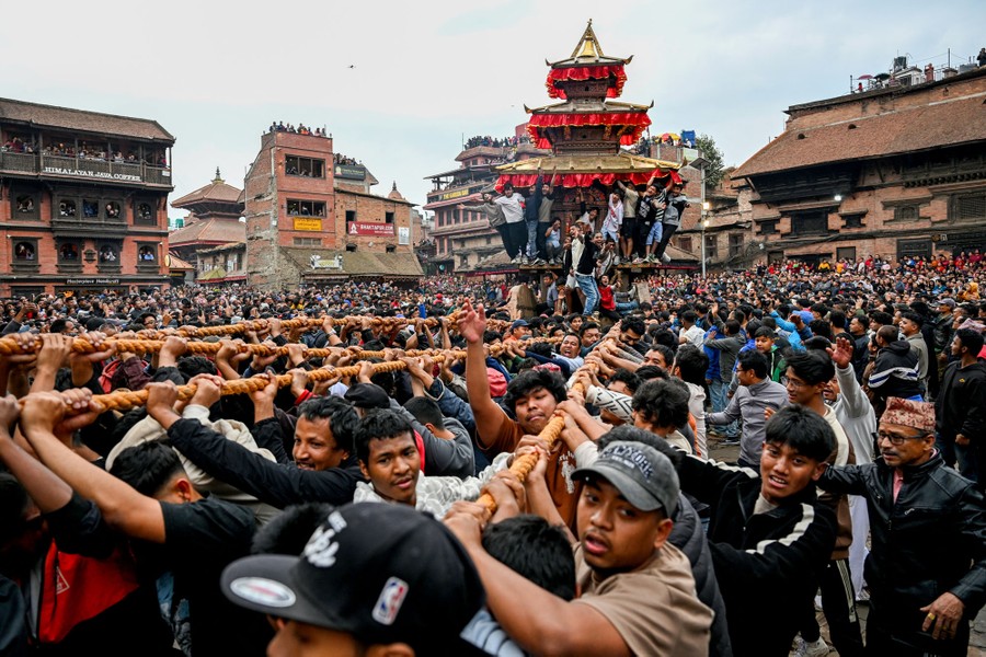 A large crowd of people gather in a city square, pulling on thick ropes to move a chariot during a festival.