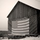 A black and white photo of a barn in the snow with a large American Flag hanging on it