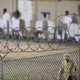 A close-up shot of a barbed-wire fence; a guard walks behind it, out of focus.