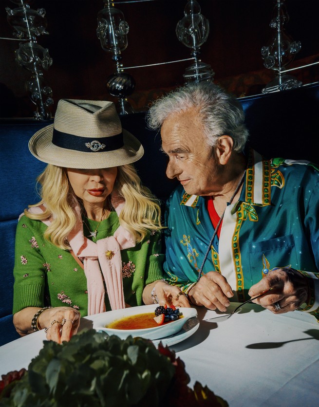 photo of blonde woman in hat and gray-haired man wearing colorful designer clothes having dessert at table