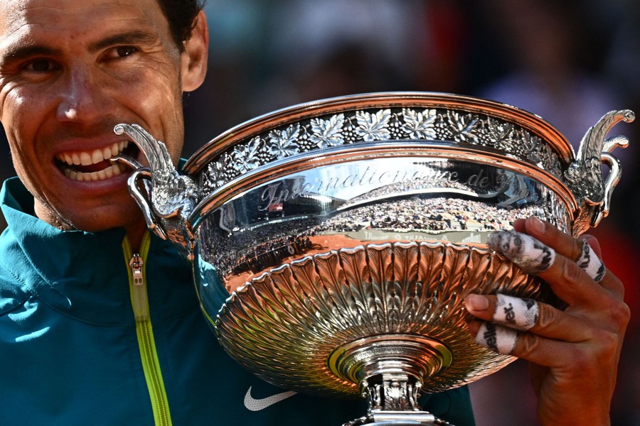 A tennis player holds a large silver trophy cup, biting one of its handles.