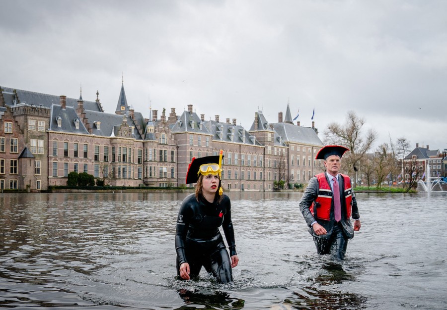Two people walk out of a shallow lake near a large building.