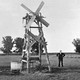 A man stands in a pasture next to a homemade windmill beside a watering trough.