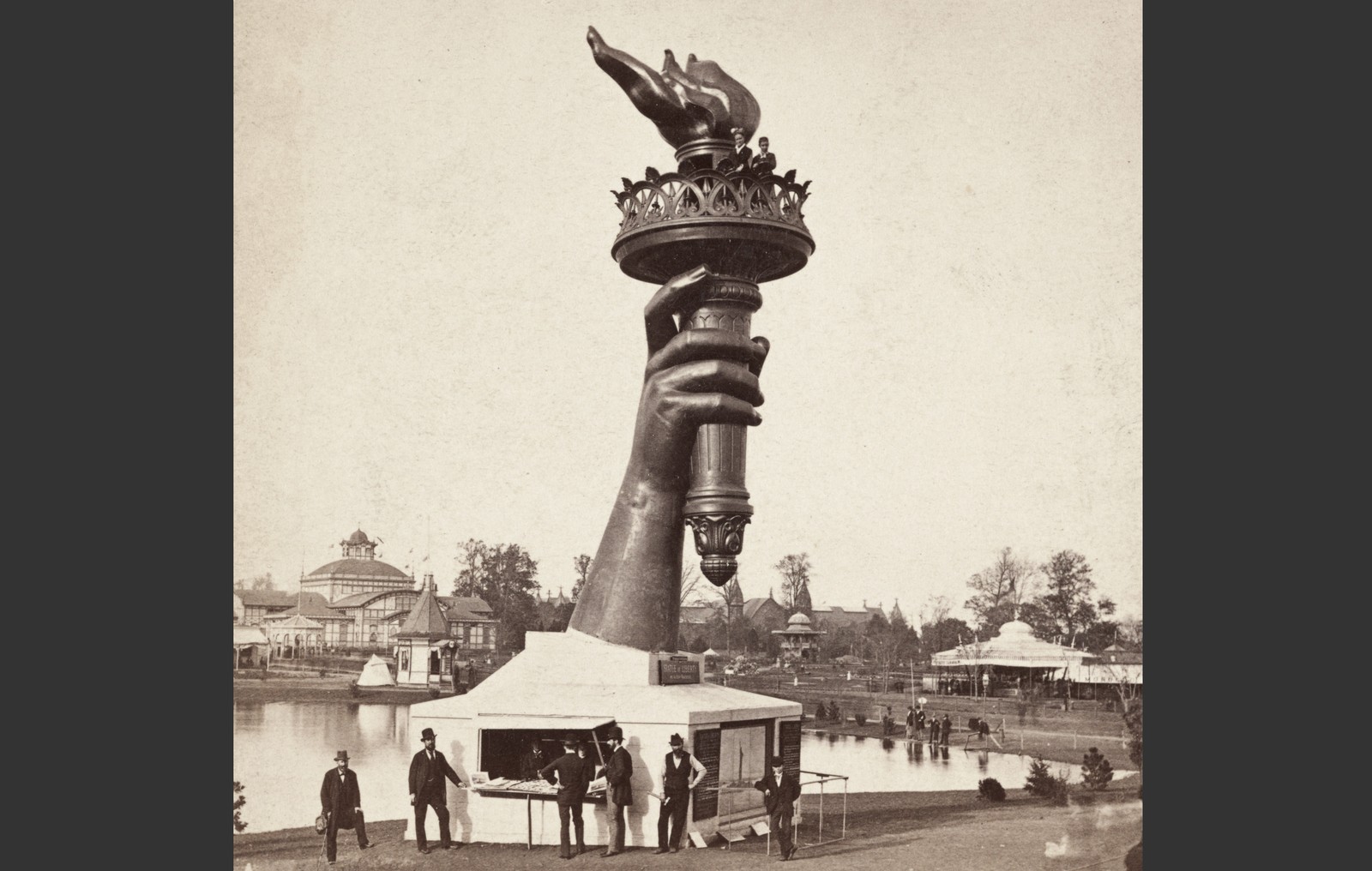 People gather at an information booth at the base of a very tall statue of an arm holding a torch, in a photo from 1876.