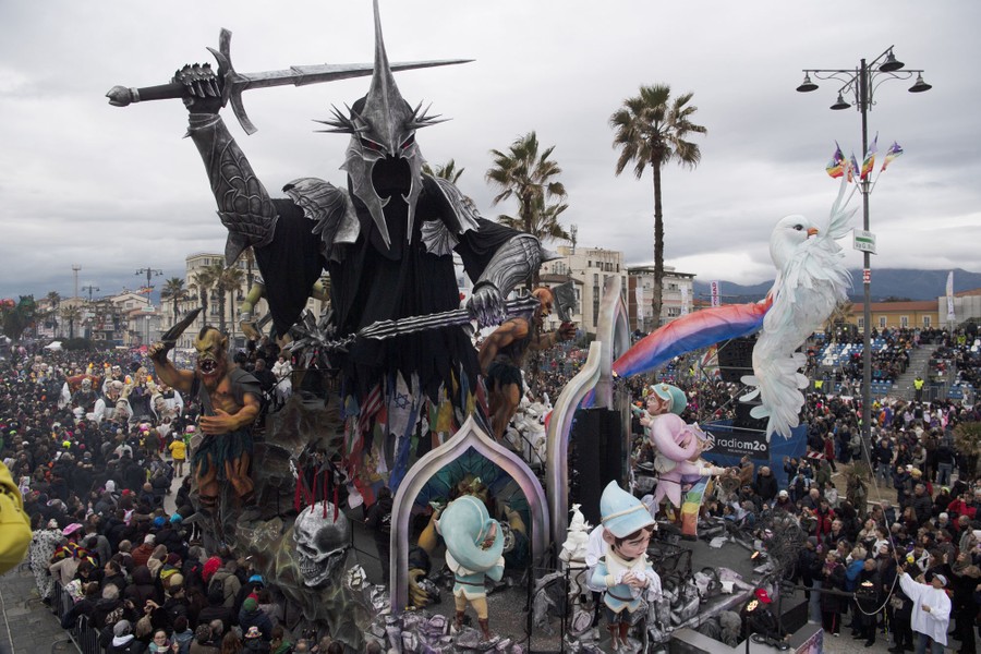A sculpture of a large frightening figure, wearing armor and wielding weapons, dominates a parade float during a carnival procession.