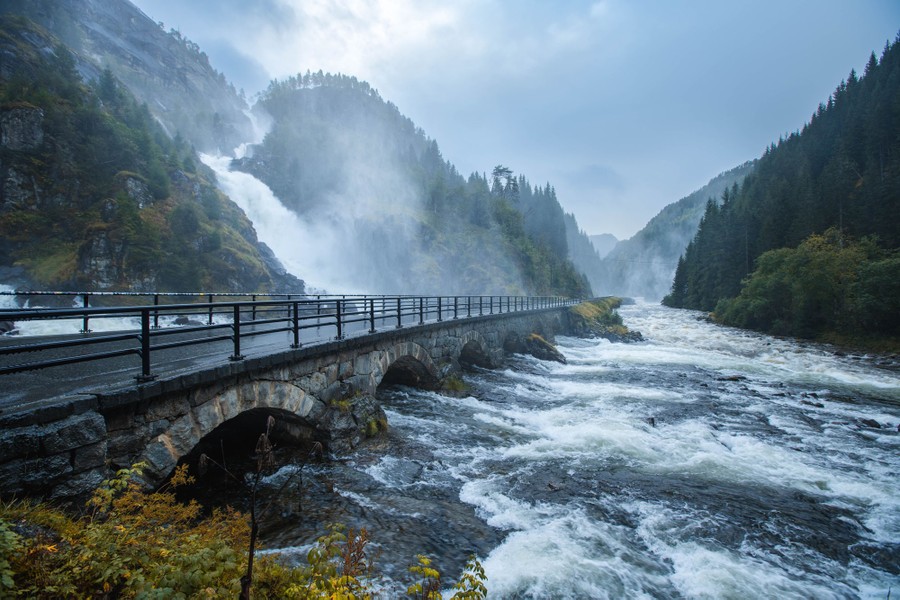 A narrow road bridge crosses in front of a waterfall, alongside a rushing river in a mountain valley.