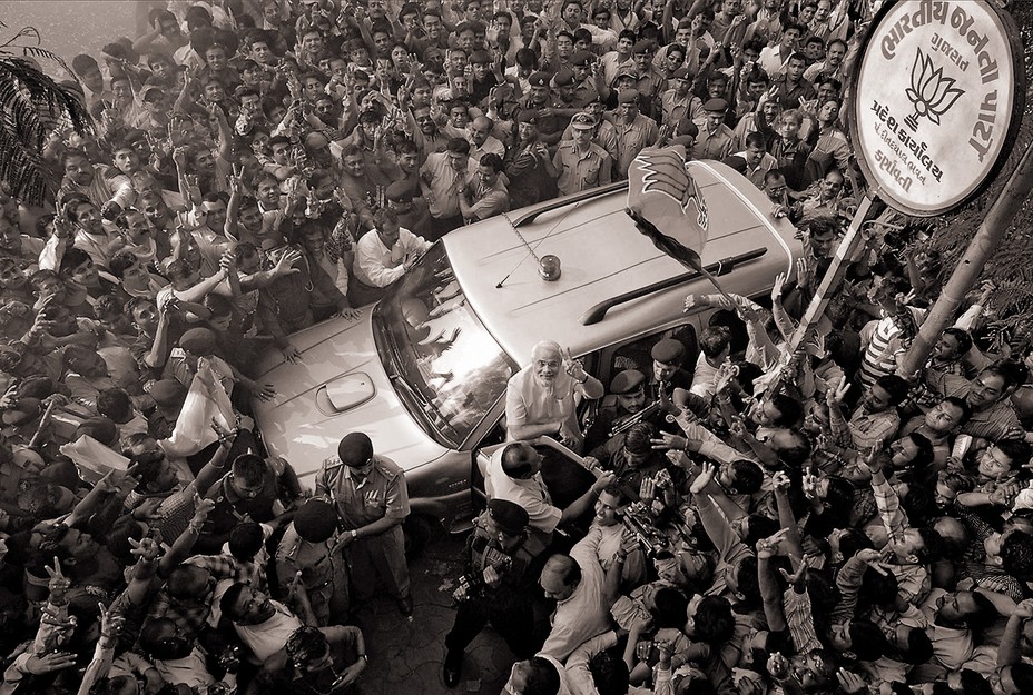 photo of Modi next to SUV surrounded by dense crowd of people, looking up and making 