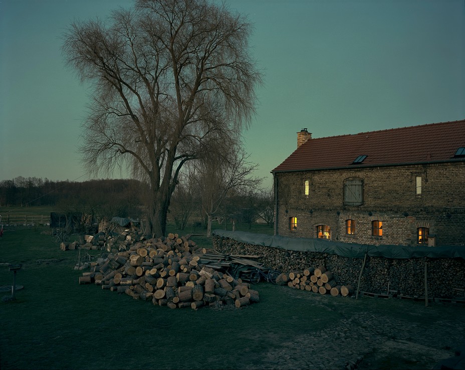 evening photograph of a woodpile and tree near a stone house with lights on in the windows