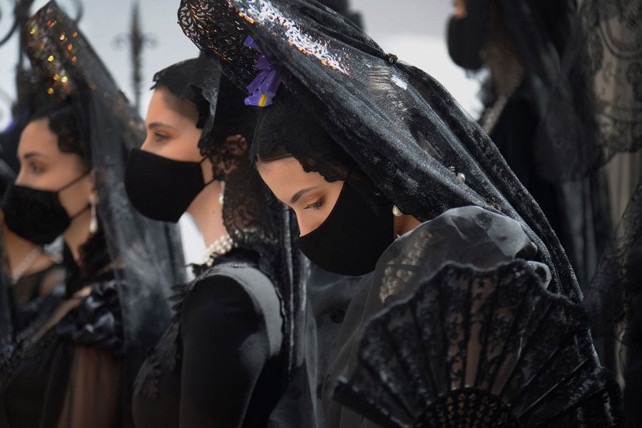 Several models gather, wearing traditional black gowns, headdresses, and protective face masks.