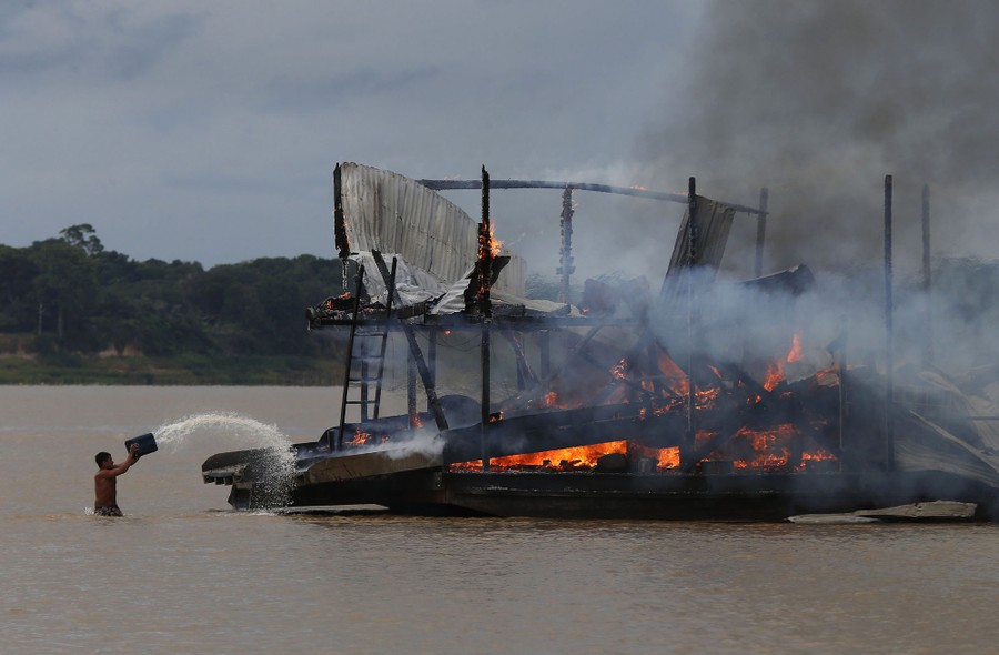 A person standing waist-deep in a river throws water on a burning barge.
